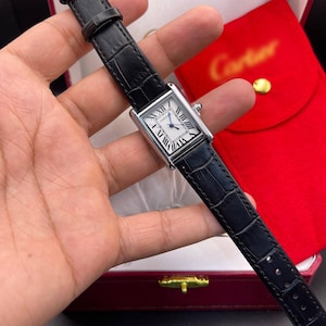 Puede incluir: Un reloj de pulsera rectangular de tono plateado con esfera blanca, números romanos negros y manecillas azules. El reloj cuenta con una correa de cuero negro texturizado que incluye una hebilla plateada y una corona plateada a juego.