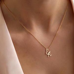 Op de afbeelding: Een delicate gouden ketting met de verweven letters "EA" als hanger. De ketting wordt getoond tegen een zachte roze achtergrond, wat het elegante ontwerp van de sieraden benadrukt. De ketting is dun en de hanger is klein, wat wijst op een minimalistische stijl.
