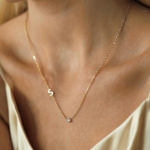 Op de afbeelding: Een delicate gouden ketting met de letter "S" en een kleine, sprankelende edelsteen hanger. De ketting wordt om de nek van een persoon gedragen, met de ketting tegen de huid. De sieraden zijn eenvoudig en elegant.