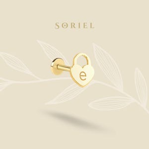 May include: A gold-colored, heart-shaped earring with the letter "e" engraved on the front. The earring has a post and a small, round backing. The word "SORIEL" is printed in a serif font above the earring.