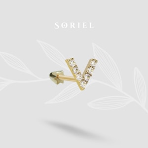 May include: A gold-colored earring in the shape of the letter "V", encrusted with small, clear gemstones. The earring has a star-shaped backing and the brand name "SORIEL" is displayed above the earring.