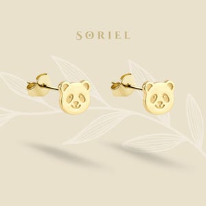 May include: A pair of gold-colored panda bear stud earrings. Each earring features a panda face with etched details. The earrings are displayed on a light beige background with the brand name "SORIEL" at the top.