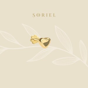 May include: A gold-colored heart-shaped earring with a flower-shaped backing. The earring is set against a neutral background with the brand name "SORIEL" in the upper center. The earring is a small, delicate piece of jewelry.