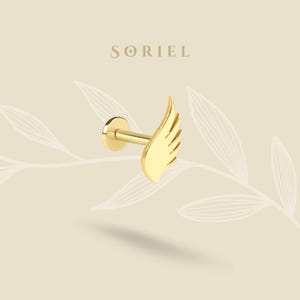 Könnte beinhalten: Ein goldfarbener Lippenpiercing-Stecker mit einem Flügeldesign. Der Stecker hat eine flache Scheibenbasis, einen geraden Stab und eine stilisierte Flügelform. Das Wort "SORIEL" ist in einer Serifenschrift über dem Stecker aufgedruckt.