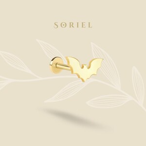 May include: A gold-coloured bat-shaped earring. The earring has a flat back and a post for piercing. The word "SORIEL" is printed in a serif font above the earring. The background is a light beige colour with a leaf design.