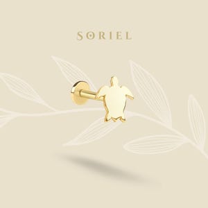 May include: A gold lip piercing with a turtle design. The piercing has a flat back and a post with a turtle-shaped charm. The word "SORIEL" is printed in a serif font above the piercing.
