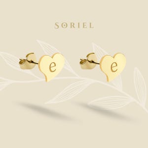 Könnte beinhalten: Ein Paar goldfarbene herzförmige Ohrstecker, jeweils mit dem Buchstaben "e" graviert. Die Ohrringe werden auf einem hellbeige Hintergrund mit dem Markennamen "SORIEL" oben angezeigt.