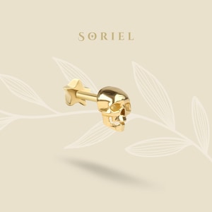 May include: A gold skull-shaped earring with a star-shaped backing. This small, detailed piece of jewellery features the brand name "SORIEL" in gold lettering above. The earring is a striking piece of body adornment.