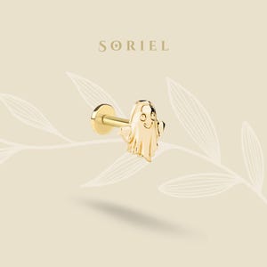 May include: A gold ghost-shaped earring with a flat back and a post. The earring features a smiling face and outstretched arms. The brand name "SORIEL" is displayed in a serif font above the earring. The background is a light beige colour with a leaf design.