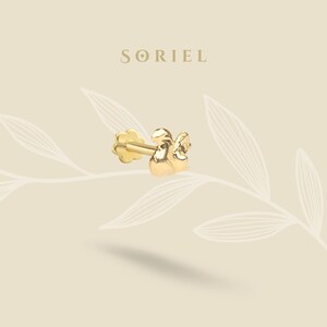 Könnte beinhalten: Goldener Ohrring mit floralem Rücken und kleinem Engel-Design. Der Ohrring befindet sich vor einem cremefarbenen Hintergrund mit dem Markennamen "SORIEL" oben.