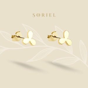以下が含まれることがあります： 三つ葉のデザインが施された、ゴールドカラーのスタッドピアス。ピアスは、繊細な葉の模様が入った淡いベージュ色の背景に展示されています。ブランド名「SORIEL」が上部に印刷されています。