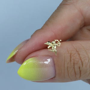 May include: A small, gold-coloured Cupid charm with a bow and arrow. The charm is in a flying pose, with wings spread. The charm is held between two fingers with a light blue background.