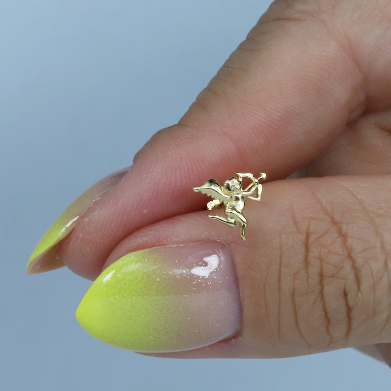 May include: A small, gold-coloured Cupid charm with a bow and arrow. The charm is in a flying pose, with wings spread. The charm is held between two fingers with a light blue background.