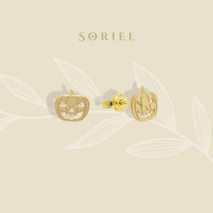 May include: Gold-colored pumpkin earrings. The earrings are in the shape of a jack-o'-lantern with carved details. The word "SORIEL" is printed in a serif font at the top of the image. The earrings are displayed on a light beige background.