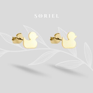 Può includere: Un paio di orecchini a bottone dorati a forma di anatra. Gli orecchini sono presentati su uno sfondo grigio chiaro, con il nome del marchio "SORIEL" in alto. Gli orecchini hanno un design semplice e minimalista.