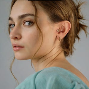May include: A close-up of a person's ear featuring gold earrings. A small stud earring is set in the upper ear, and a simple gold hoop earring is in the lobe. The person is wearing a light blue top.