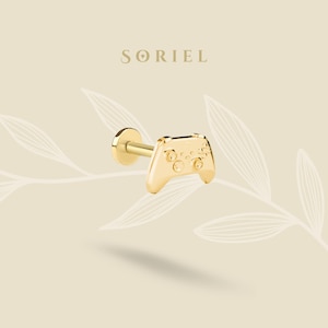 Puede incluir: Un pendiente de botón de color dorado con forma de mando de videojuego. El pendiente tiene una parte trasera plana y un poste para perforar. El diseño del mando incluye botones y un panel direccional. La marca "SORIEL" se muestra encima del pendiente.