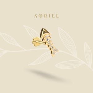Pode incluir: Um brinco dourado em forma de esqueleto de peixe, adornado com pequenas pedras preciosas transparentes. O brinco está sobre um fundo neutro com o nome da marca "SORIEL" no topo.
