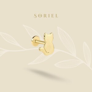 May include: A gold-colored cat-shaped earring stud. The earring features a flat, round backing and a post. The cat silhouette is sitting with its tail curled. The brand name "SORIEL" is displayed in a serif font.