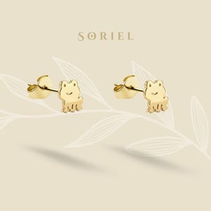 May include: A pair of gold-colored frog stud earrings. Each earring features a small, smiling frog design. The earrings are displayed on a light beige background with the brand name "SORIEL" at the top.
