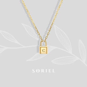 May include: A gold-coloured necklace with a padlock pendant, engraved with the letter "C". The chain is delicate, and the pendant is square. The necklace is set against a light grey background with a floral design and the brand name "SORIEL".