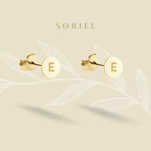 Puede incluir: Un par de pendientes de botón dorados, cada uno con un diseño circular y la letra "E" en el centro. Los pendientes se muestran sobre un fondo beige claro con un sutil patrón floral y el nombre de la marca "SORIEL" en la parte superior.