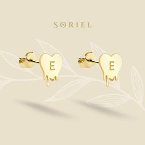 May include: Gold-coloured heart-shaped stud earrings featuring the letter "E" and a dripping design. The earrings are presented on a light beige background with the brand name "SORIEL" at the top.