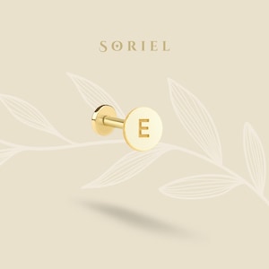 May include: A gold-colored stud earring with the letter "E" engraved on a circular disc. The earring has a post and a flat back. The brand name "SORIEL" is displayed in gold lettering at the top of the image. The background is a light beige color with leaf-like designs.