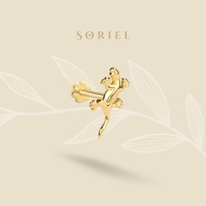 Può includere: Un orecchino a forma di lucertola color oro, con un perno e un retro a forma di fiore. L'orecchino è posizionato in diagonale su uno sfondo beige chiaro con il marchio "SORIEL" in alto.