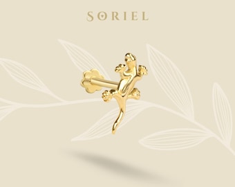 May include: A gold-colored lizard-shaped earring with a post and a flower-shaped backing. The earring is positioned diagonally against a light beige background with the brand name "SORIEL" at the top.