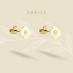May include: A pair of gold stud earrings, each with a four-leaf clover design and the letter "e" in the centre. The earrings are set against a light beige backdrop, with the brand name "SORIEL" displayed above them.