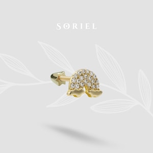 May include: A gold-colored earring featuring a rainbow design with clear, sparkling stones. The earring has a star-shaped backing and cloud-shaped ends. The brand name "SORIEL" is displayed in the upper center.