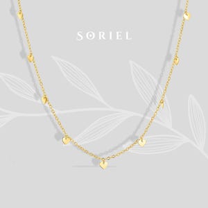 Pode incluir: Um delicado colar dourado com pequenos pingentes em forma de coração. O colar é suspenso por uma corrente fina e é exibido em um fundo cinza claro com o nome da marca "SORIEL" no centro superior.