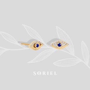 Puede incluir: Pendientes de botón dorados con diseño de ojo. Cada pendiente presenta un centro azul y está rodeado de pequeñas piedras transparentes. Los pendientes se muestran sobre un fondo gris claro con un diseño de hojas. La marca "SORIEL" está en la parte inferior.