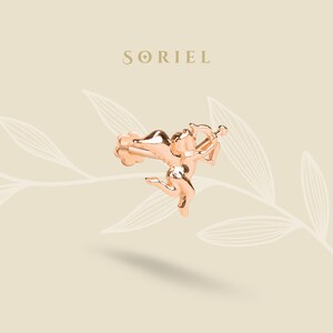 May include: A rose gold Cupid charm, with a bow and arrow, in mid-flight. A cloud-like shape is behind the charm. The word "SORIEL" is above the charm in gold. The charm is a decorative item.