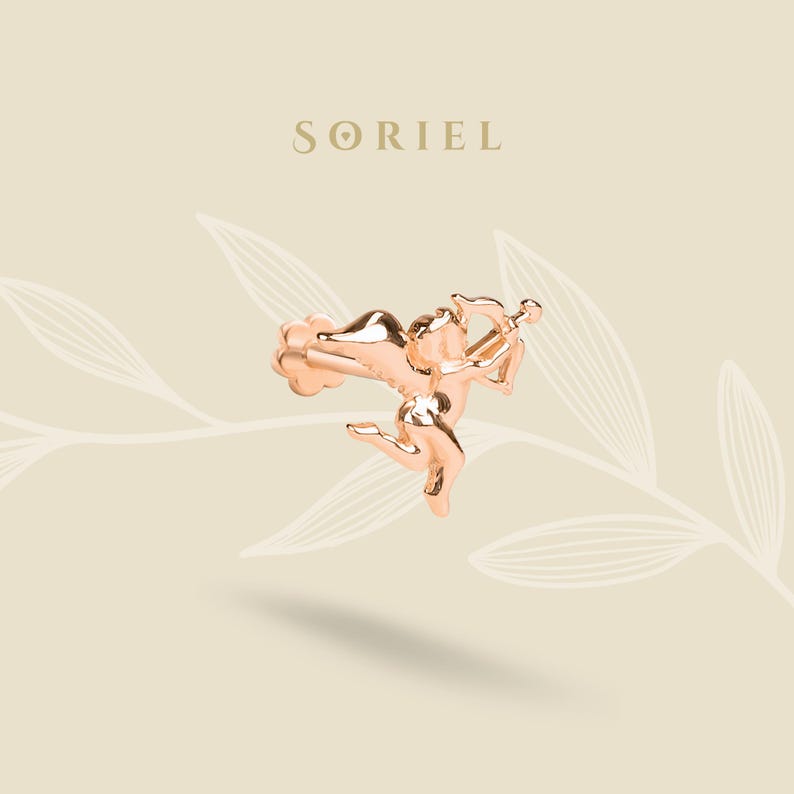 May include: A rose gold Cupid charm, with a bow and arrow, in mid-flight. A cloud-like shape is behind the charm. The word "SORIEL" is above the charm in gold. The charm is a decorative item.