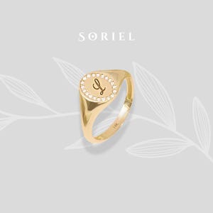 Könnte beinhalten: Ein goldener Siegelring mit einem ovalen, mit Diamanten besetzten Gesicht. Das Gesicht zeigt den Buchstaben "L" in einer Schreibschrift. Der Ring ist mit "14K" gestempelt und der Markenname "SORIEL" wird darüber angezeigt.