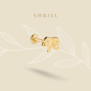Könnte beinhalten: Goldfarbener Ohrstecker mit Elefantenmotiv. Der Ohrring hat einen Stecker und eine flache, runde Rückseite. Das Wort "SORIEL" ist in einer serifenbetonten Schriftart über dem Ohrring aufgedruckt. Der Hintergrund ist hellbeige mit einem Blattmuster.