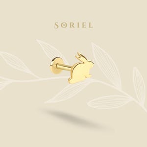 Könnte beinhalten: Ein goldfarbener Lippenpiercing-Stecker mit einer Kaninchensilhouette. Der Stecker hat eine flache, kreisförmige Rückseite und einen geraden Stab. Der Markenname "SORIEL" ist in einer Serifenschrift über dem Stecker abgebildet. Der Hintergrund ist hellbeige mit einem Blattdesign.