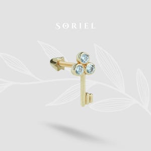 May include: A gold-colored key-shaped earring with three light blue gemstones. The earring has a star-shaped backing. The word "SORIEL" is printed in the upper center of the image.