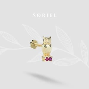 Pode incluir: Um brinco dourado em forma de coruja com três pequenas pedras vermelhas na base. O brinco tem uma superfície texturizada e está sobre um fundo neutro e suave. A marca "SORIEL" é exibida na parte superior.