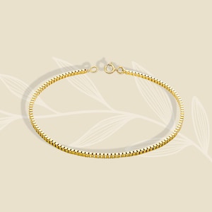 Op de afbeelding: Een goudkleurige box chain armband bestaande uit een reeks onderling verbonden vierkante schakels die een gladde, doorlopende band vormen. De armband is aan de uiteinden voorzien van een kleine, ronde veerringsluiting voor een strak uiterlijk.