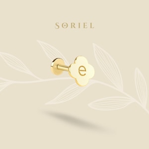 May include: A gold-colored earring with a four-leaf clover design and the letter "e" engraved in the center. The earring has a flat back and a post for piercing. The word "SORIEL" is above the earring.