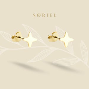 Puede incluir: Un par de pendientes de botón dorados con forma de estrella. Los pendientes tienen un diseño de estrella de cuatro puntas y están sobre un fondo beige claro con el nombre de la marca "SORIEL" en la parte superior.