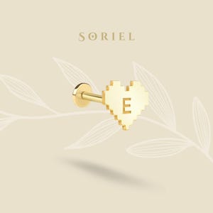 May include: A gold stud earring with a pixelated heart design and the letter "E". The earring has a flat back and a post. The brand name "SORIEL" is displayed at the top. The background is a light beige color.