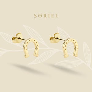 Könnte beinhalten: Ein Paar goldfarbene Hufeisen-Ohrstecker. Die Ohrringe sind vor einem hellbeige Hintergrund mit dem Markennamen "SORIEL" oben platziert. Das Hufeisen-Design ist ein klassisches Glückssymbol.