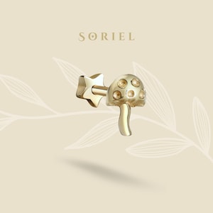 Puede incluir: Pendiente dorado con forma de seta y cierre en forma de estrella. El sombrero de la seta presenta pequeños detalles redondos y claros. El pendiente está sobre un fondo neutro con la marca "SORIEL" en la parte superior.