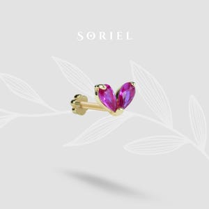Puede incluir: Pendiente dorado con un poste y una cabeza decorativa con dos piedras preciosas de color magenta, talla marquesa. La palabra "SORIEL" está impresa en una fuente gris claro encima del pendiente.