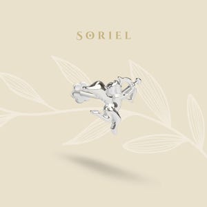 May include: A silver earring in the shape of Cupid, complete with a bow and arrow. The earring is intricately designed, with the word "SORIEL" in a serif font above. The background is a light beige with a subtle leaf pattern.