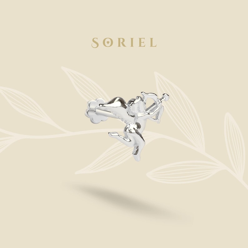 May include: A silver earring in the shape of Cupid, complete with a bow and arrow. The earring is intricately designed, with the word "SORIEL" in a serif font above. The background is a light beige with a subtle leaf pattern.